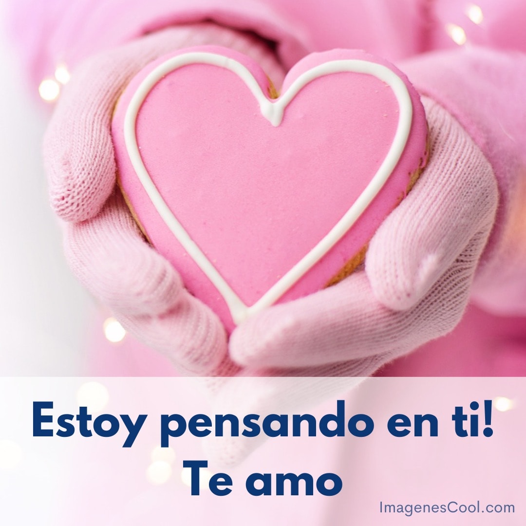 manos con guantes sostienen galleta en forma de corazón rosa. texto: estoy pensando en ti! te amo